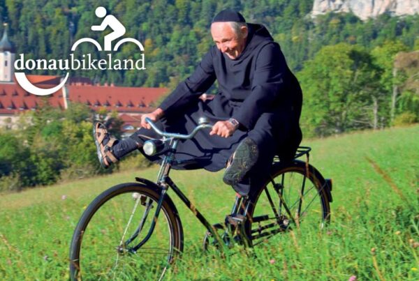 Donaubergland Postkartenmotiv mit fahrradfahrendem Mönch, der die Beine in die Luft hält und oberhalb eines Klosters in der Wiese fährt.