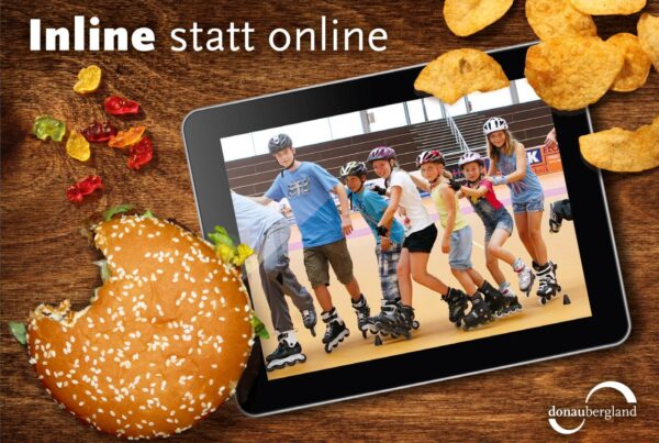 Donaubergland Postkartenmotiv mit Tablet auf Tisch mit Inline-Fahrern auf Bildschirm und angebissener Burger, Gummibärchen und Chips auf dem Tisch.