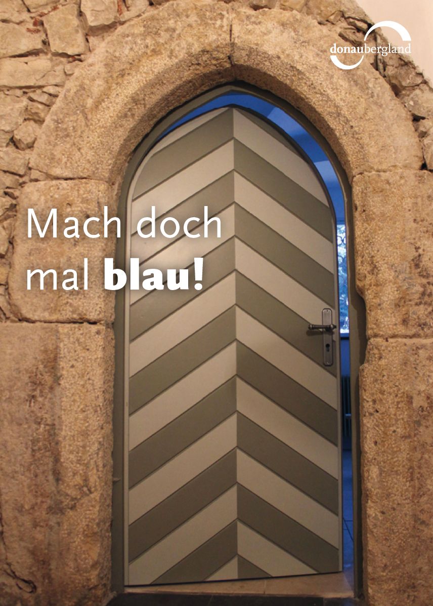 Donaubergland Postkartenmotiv mit Tür in einer antiken Mauer, die einen Spalt weit geöffnet ist und den schmalen Blick in eine blaues Zimmer freigibt.