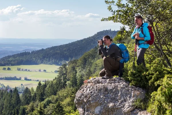 Bild von zwei Wanderern, der eine sitzt auf einem Felsen und fotografiert mit einer Kamera und der andere steht dahinter mit Wanderstöcken